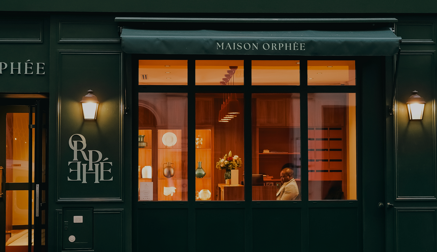 Façade d'une boutique nommée Maison Orphée avec une vitrine éclairée montrant un homme assis et des décorations visibles à l'intérieur.