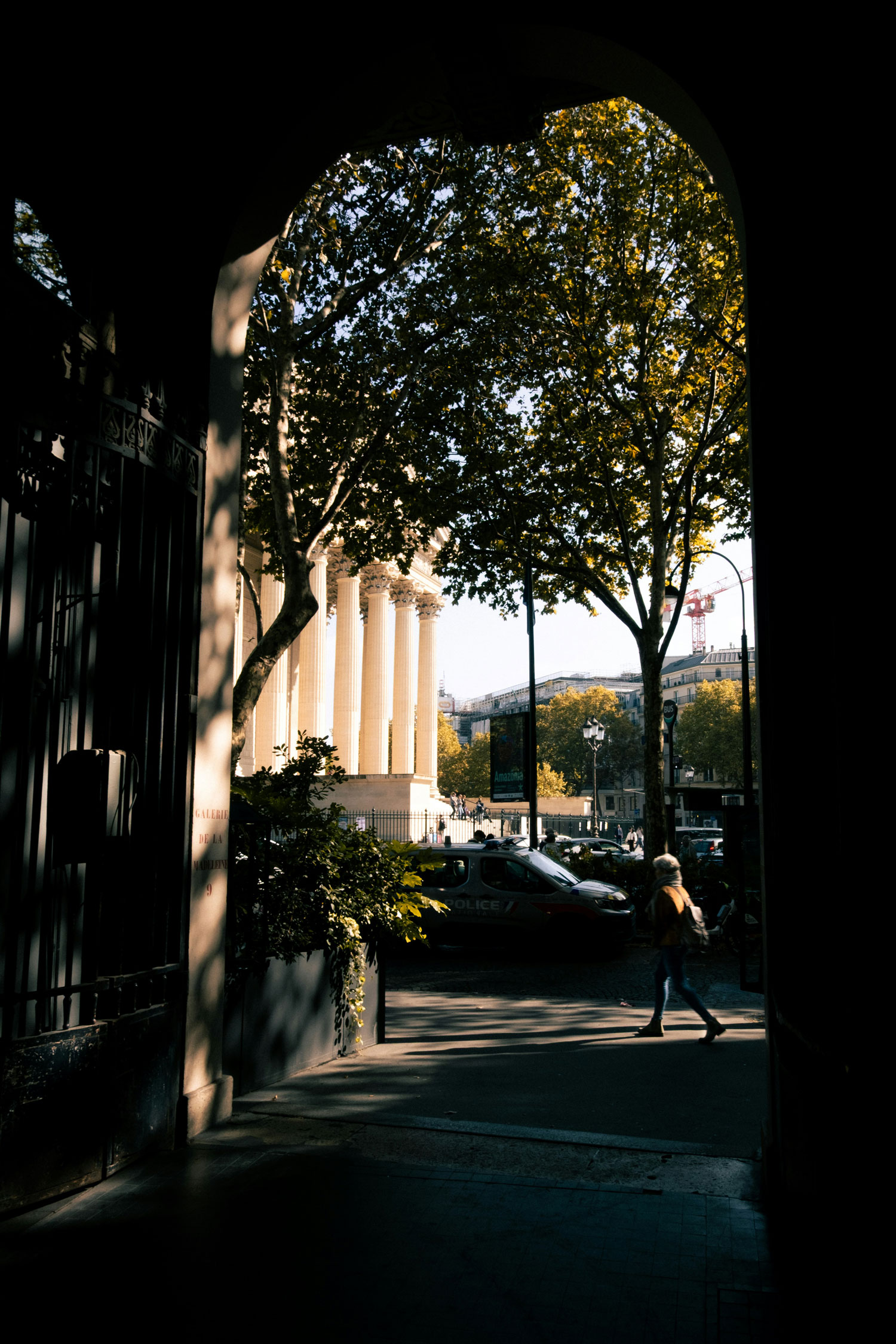 Persona che cammina sotto un arco verso un edificio con grandi colonne bianche, alberi e un'auto della polizia parcheggiata.
