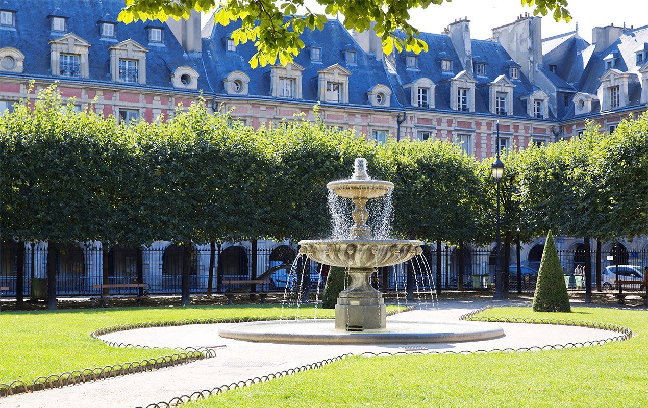 Fontaine à deux niveaux avec de l'eau s'écoulant, entourée d'herbe et d'arbres taillés, avec un bâtiment historique en arrière-plan.
