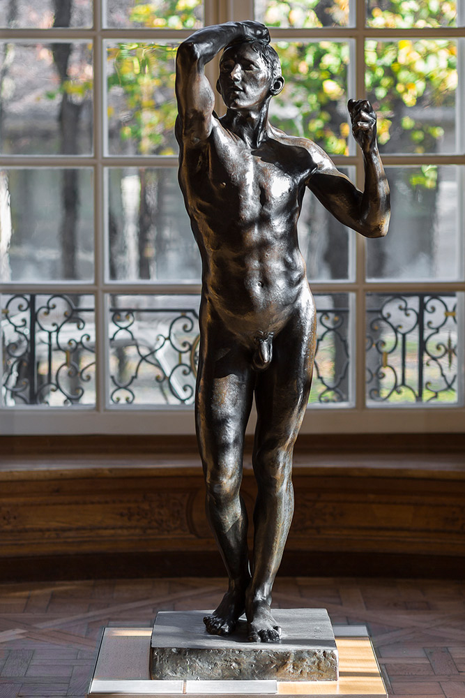 Statue en bronze d'un homme nu debout, une main sur le front et l'autre levée en poing, devant une grande fenêtre avec barreaux décoratifs.