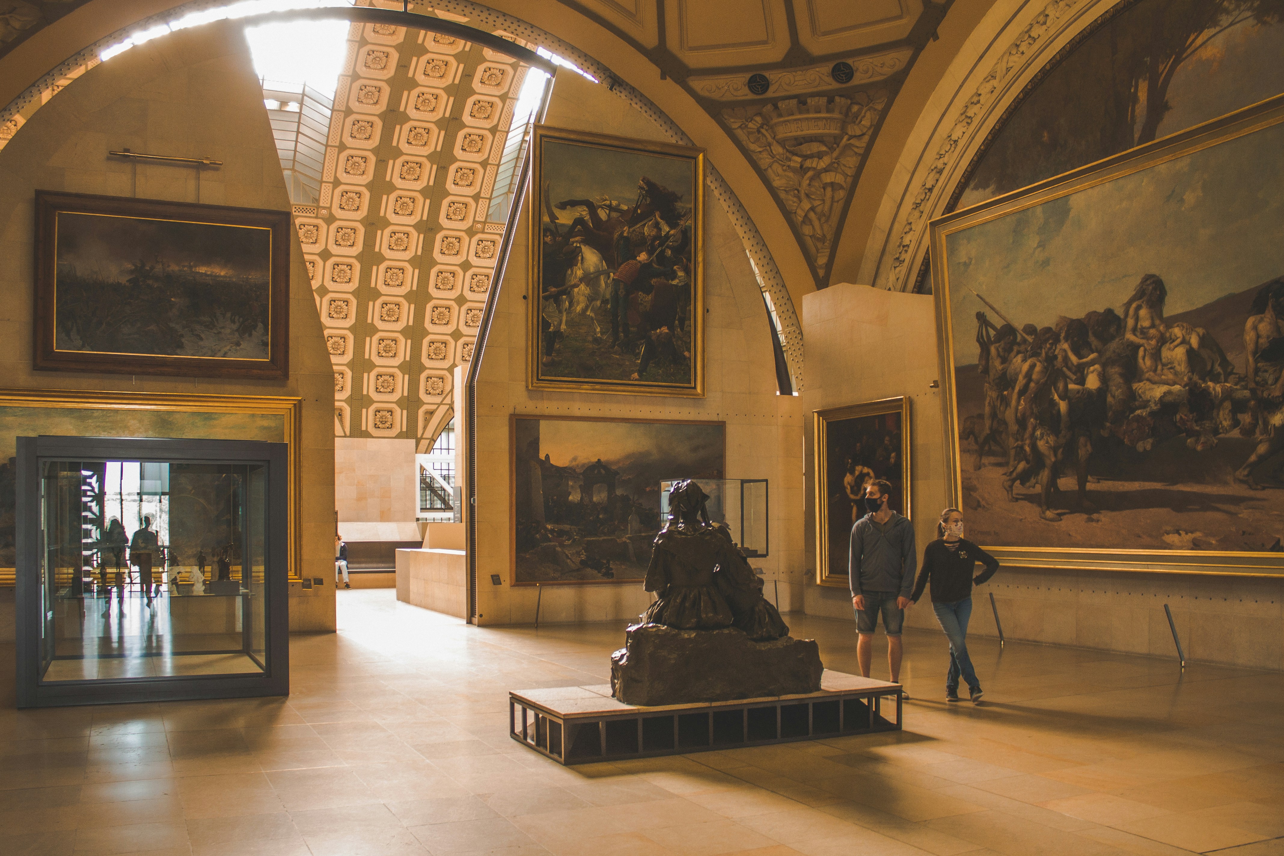Intérieur d'une galerie d'art avec de grandes peintures murales, une statue au centre et deux personnes masquées tenant la main en train de regarder les œuvres.