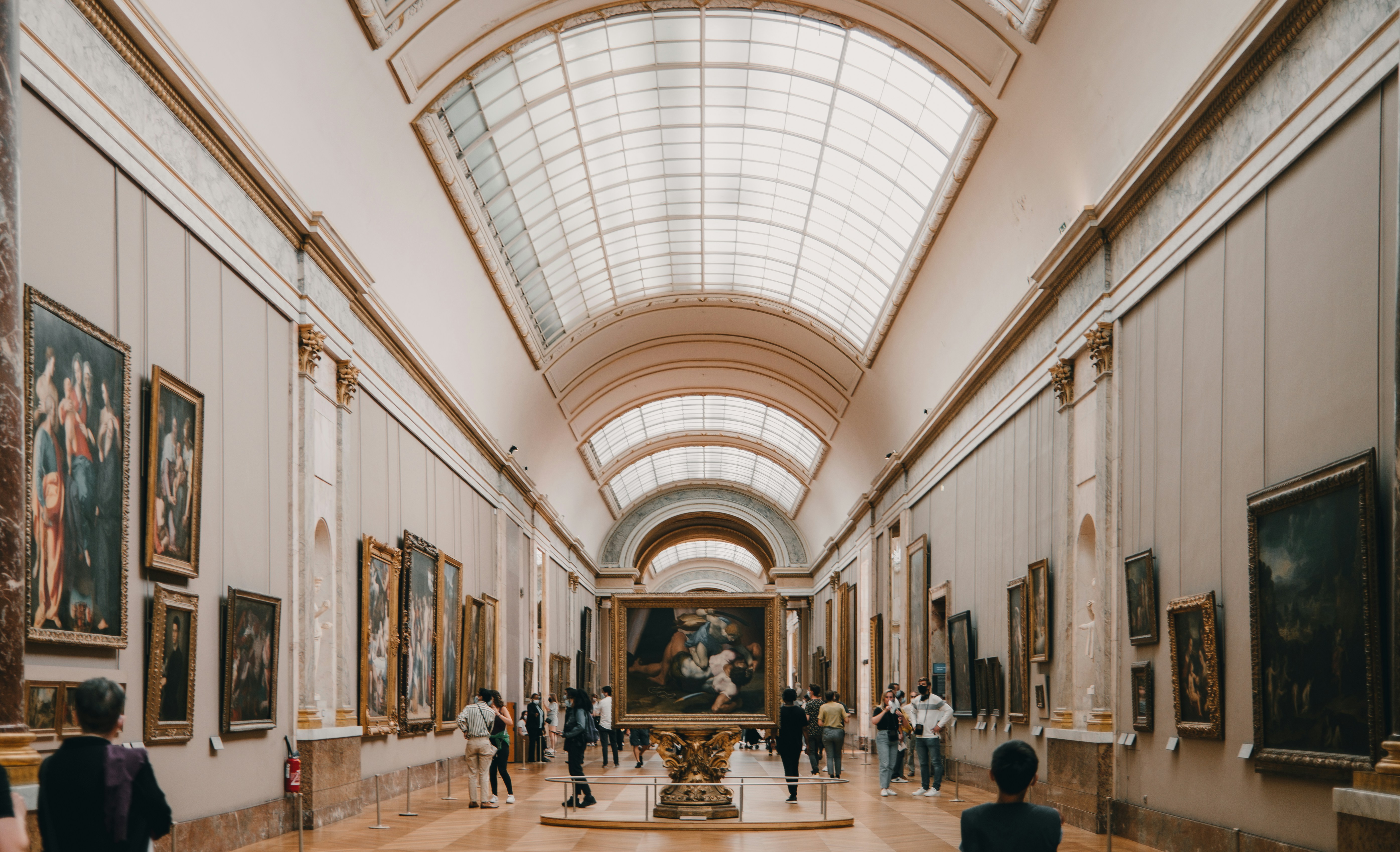 Salle de musée avec un plafond voûté en verre et plusieurs peintures encadrées exposées aux murs, des visiteurs regardent les œuvres.