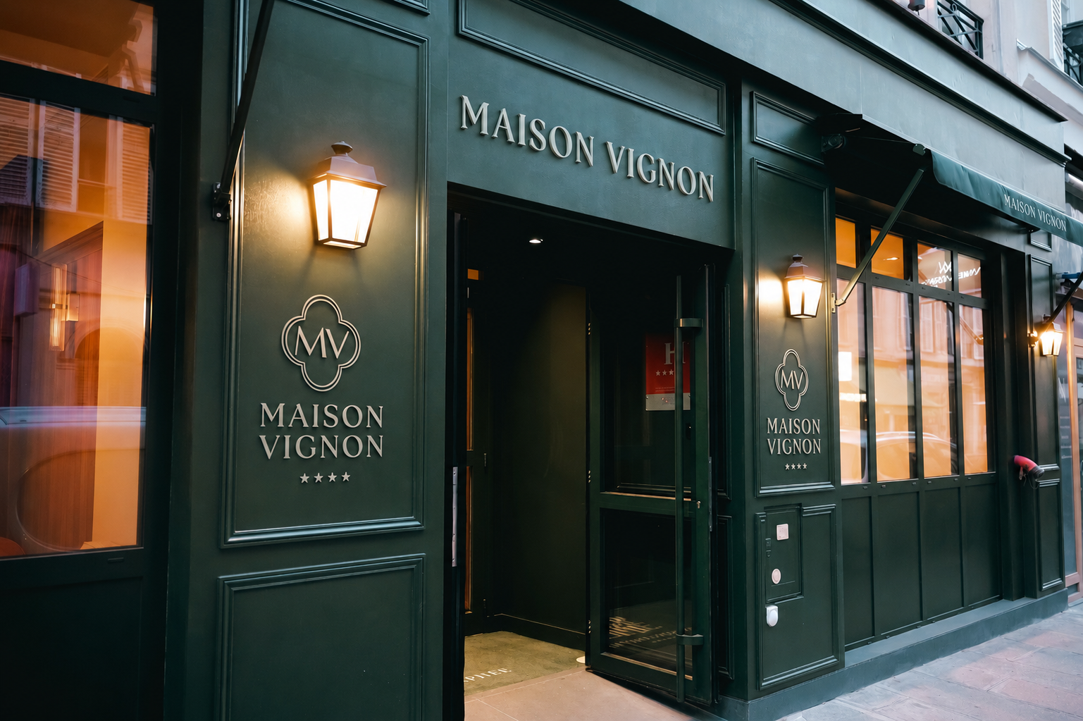 Façade sombre avec enseigne 'MAISON Vignon', vitrine éclairée montrant un homme assis à un bureau et des étagères avec des objets décoratifs.