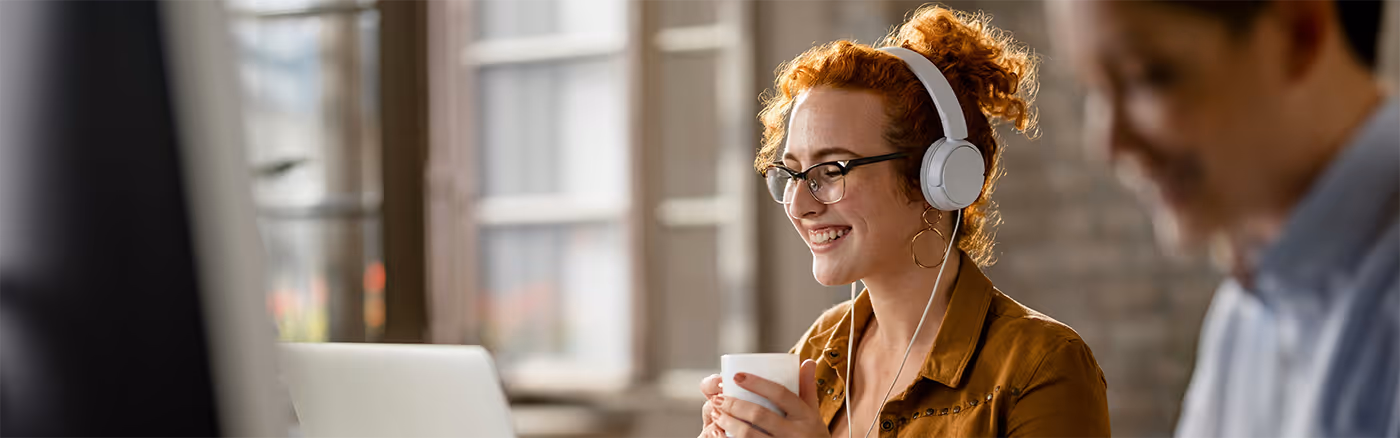 Vrouw met rode krullen, zwarte bril en witte koptelefoon die lacht en een kopje vasthoudt terwijl ze voor een laptop zit.