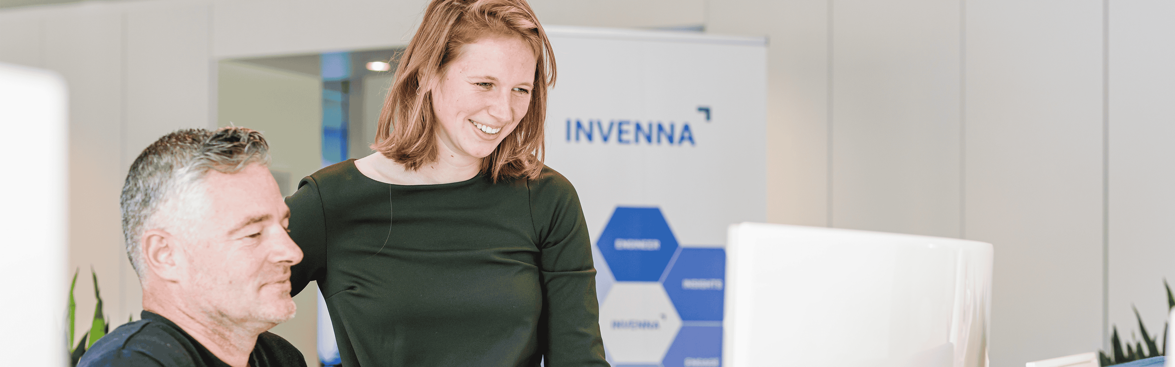 Twee collega's bespreken iets lachend bij een computer in een kantoorruimte met een Invenna-bord op de achtergrond.