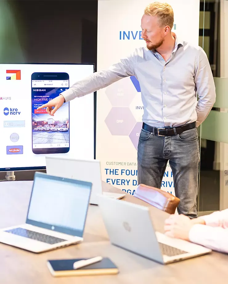 Man wijst naar scherm met smartphone display tijdens presentatie in kantoorruimte met laptops op tafel.