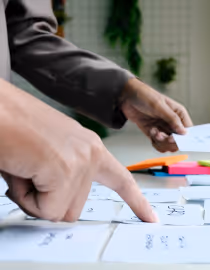 Hand wijst op papieren met aantekeningen verspreid op een tafel, achtergrond met planten.