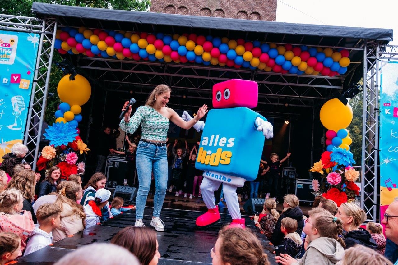 Children and adults interacting with a person in a colorful block costume at an outdoor event, with one adult wearing a pink shirt that says 'alles kids' and an inflatable red backdrop.