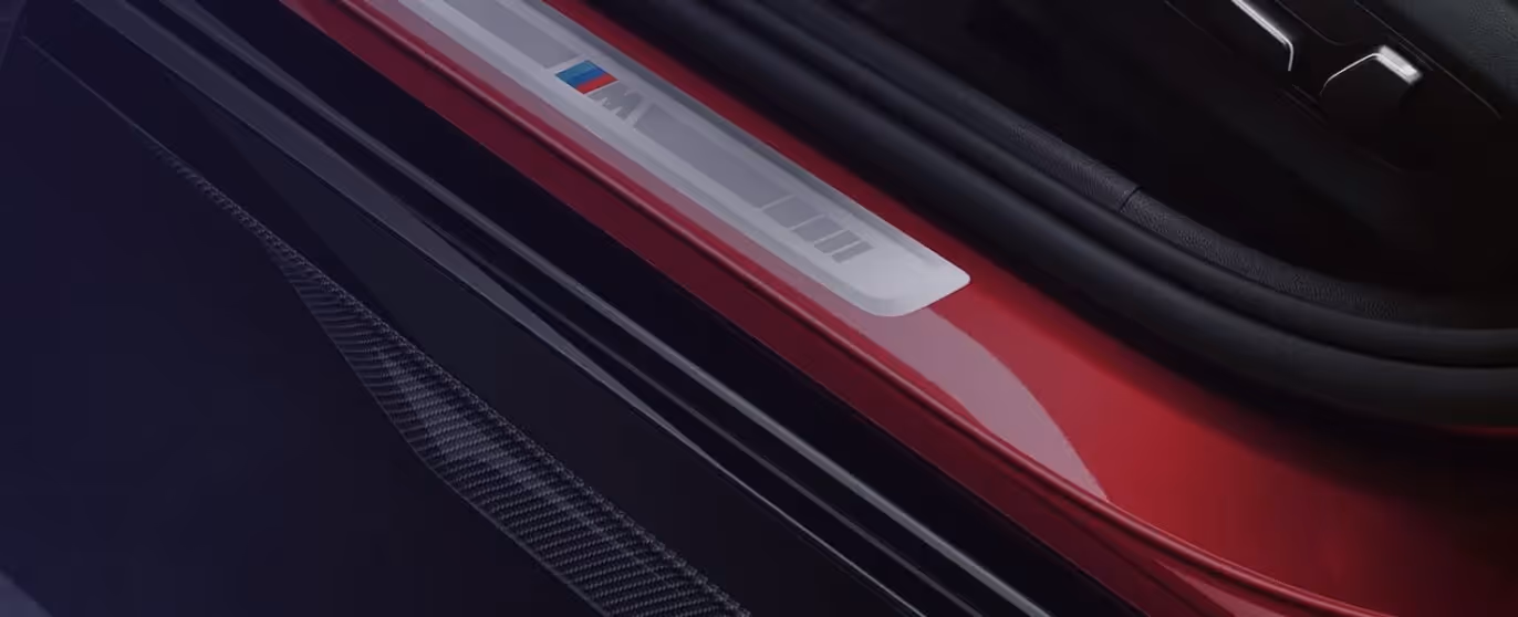 Close-up view of a car door sill with a BMW M logo plaque and red exterior paint.