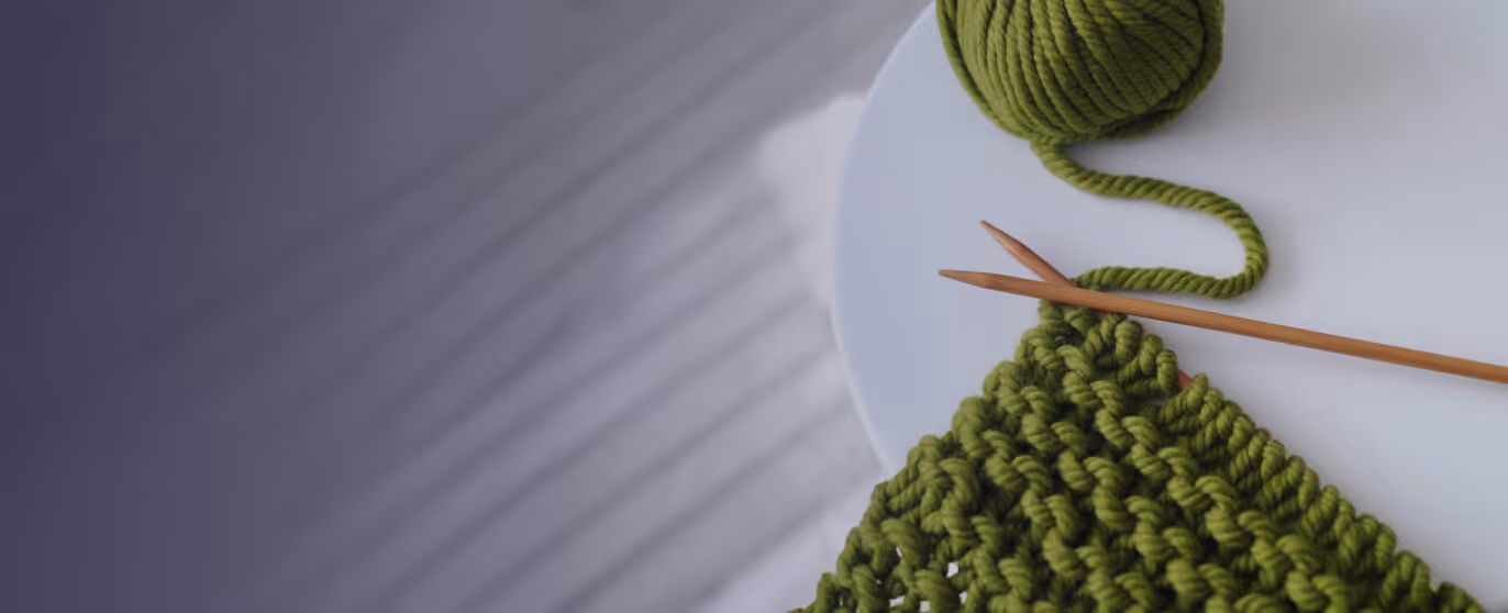 Close-up of green yarn ball and knitted green wool with wooden knitting needles on a white surface.