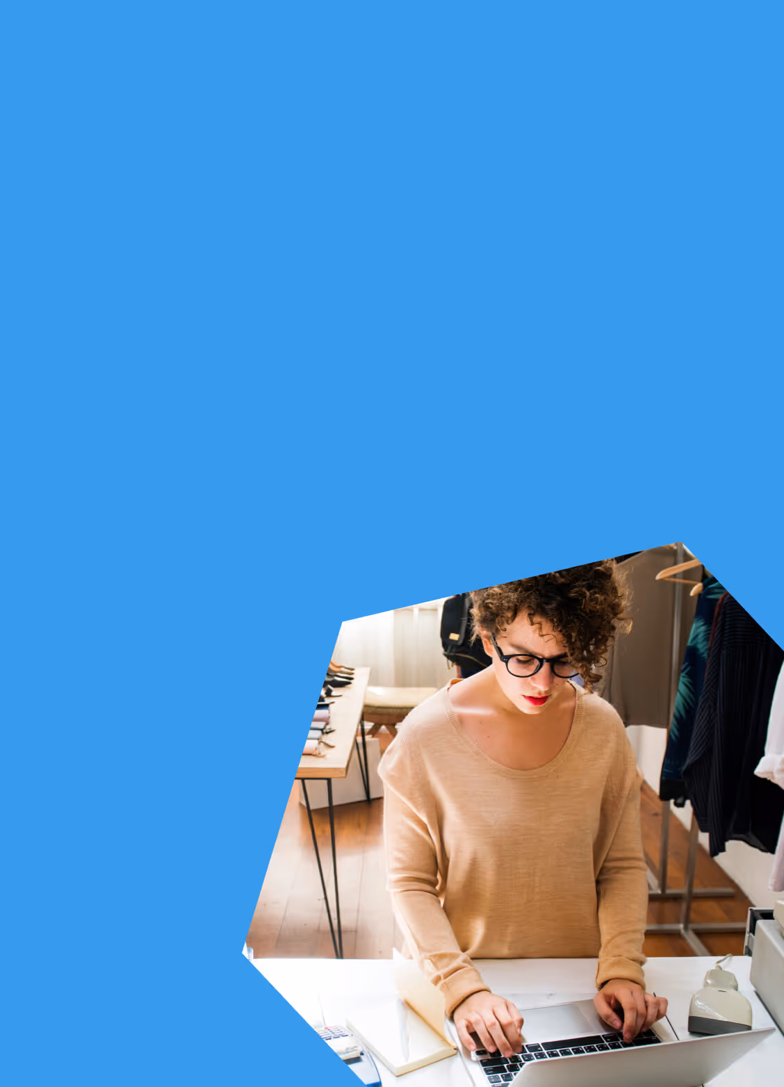 Person with curly hair and glasses working on a laptop at a desk in a clothing store.