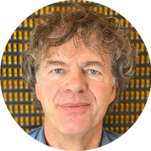 Close-up portrait of a middle-aged man with curly hair and blue eyes smiling slightly against a patterned yellow and black background.
