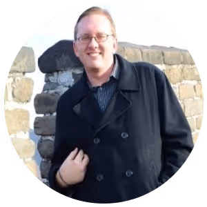 Person wearing glasses and a black coat standing outdoors in front of a stone wall, smiling.