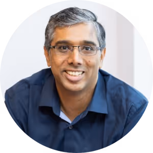 Smiling man with short gray hair and glasses wearing a blue collared shirt against a white background.