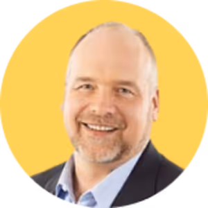 Smiling middle-aged man with a light beard and mustache, wearing a dark blazer and light blue shirt, against a yellow circular background.