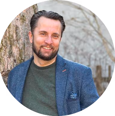 Smiling man with beard wearing a blue blazer and green sweater standing outdoors by a tree.