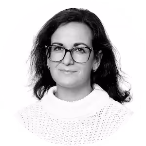 Woman with dark hair wearing glasses and a white sweater, smiling slightly against a plain background.