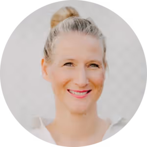 Portrait of a smiling woman with blonde hair in a bun wearing a white top against a neutral background.
