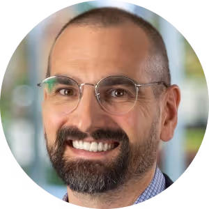 Smiling man with glasses, short dark hair and beard, wearing a blue checkered shirt with blurred background.