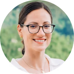 Smiling woman with dark hair pulled back, wearing glasses and gold hoop earrings, with a blurred green background.