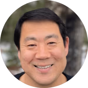 Smiling man with short black hair and a blurred outdoor background.