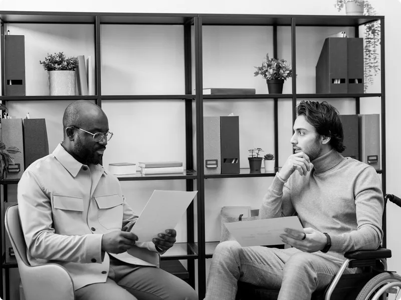 Two men sitting and reviewing documents in an office, one in a wheelchair and the other in a chair.