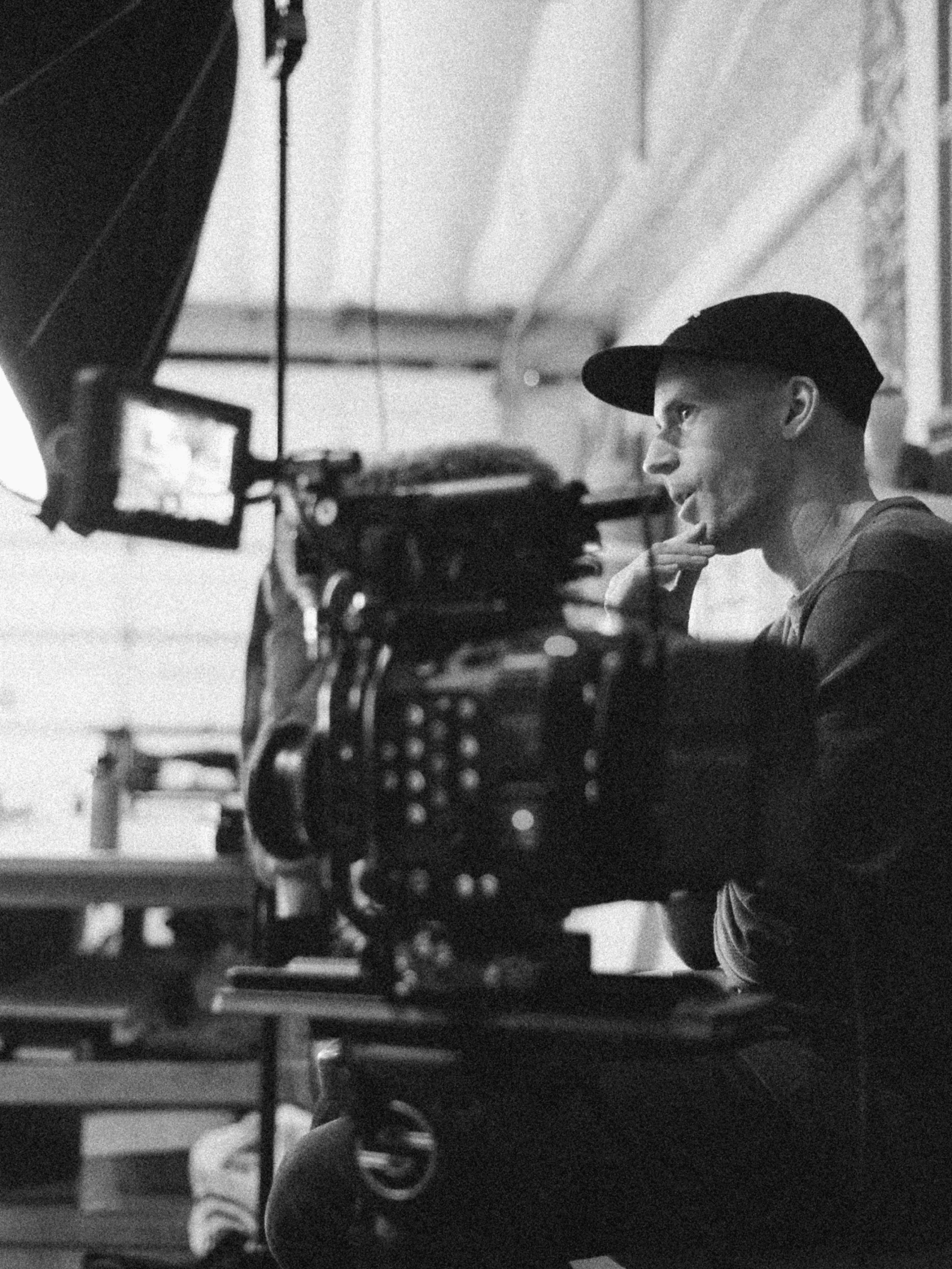 A person in a cap sits next to professional video equipment, appearing thoughtful. The scene is shot in black and white, with a camera and lighting setup visible in the foreground.