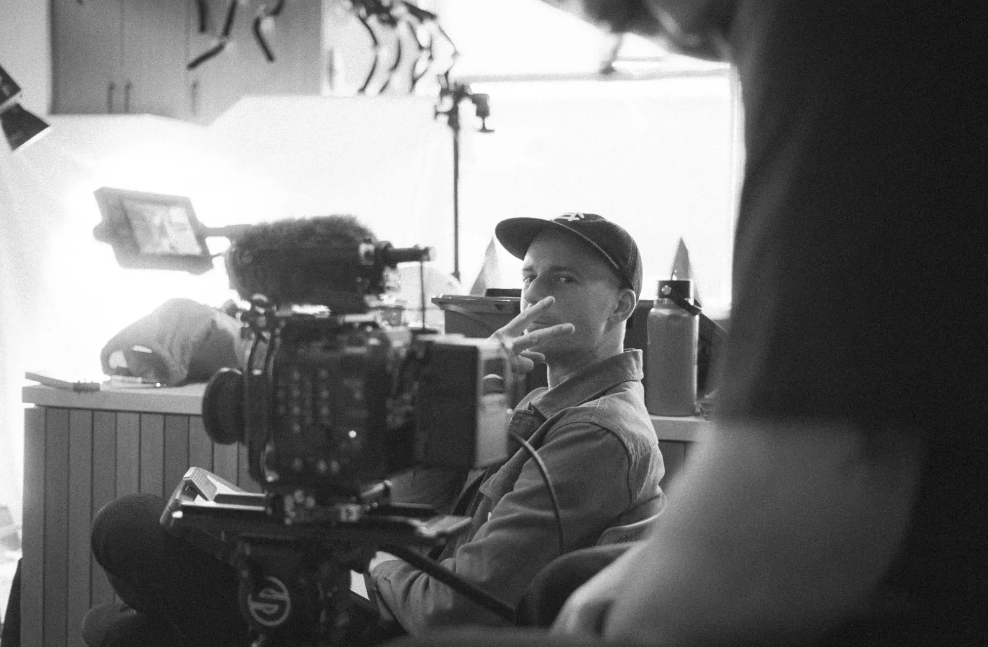 A person in a cap sits behind a professional video camera, making a peace sign with their hand. The scene appears to be on a film set, with equipment and another person out of focus in the foreground.