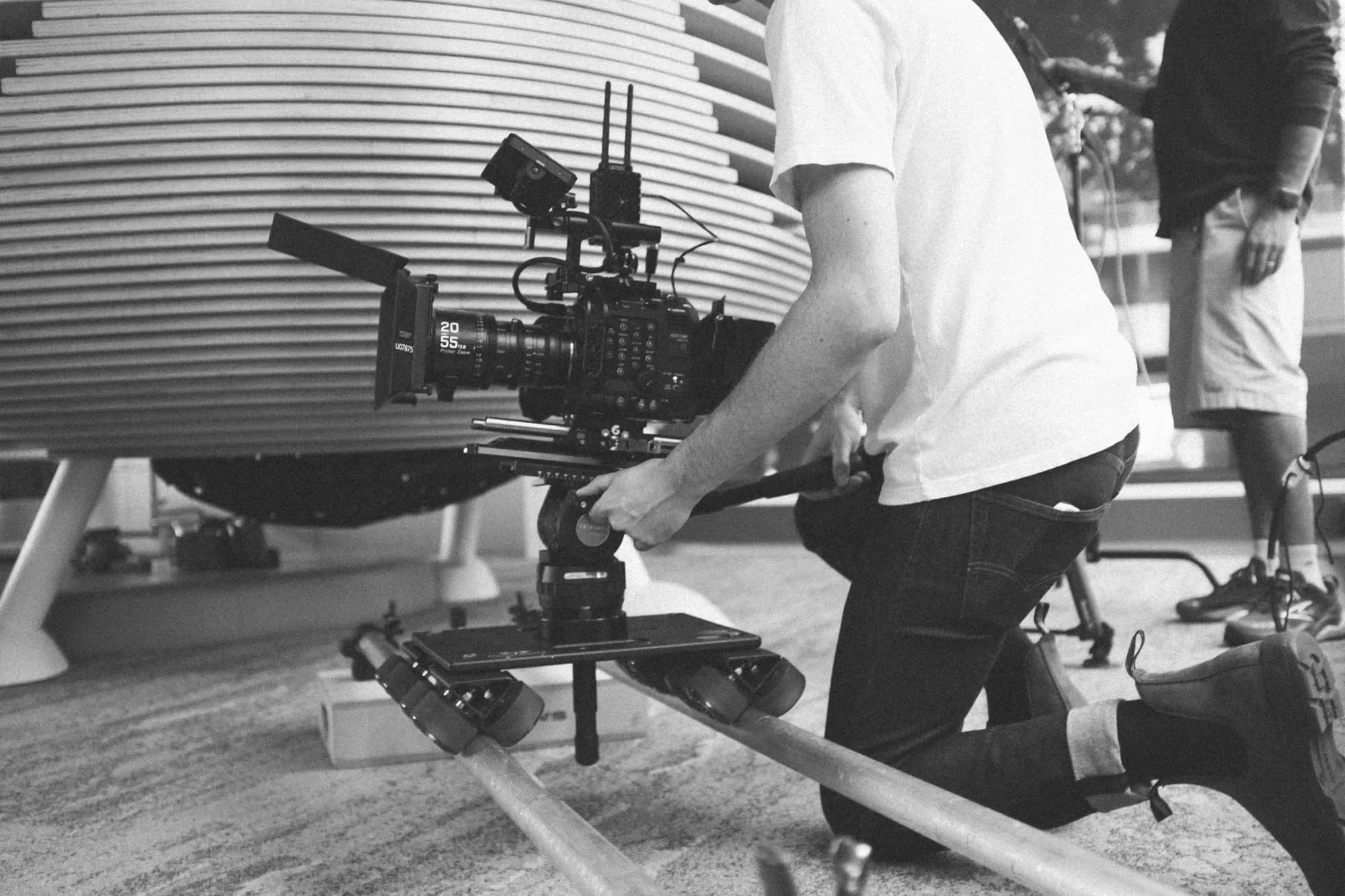 A person kneels on the ground, operating a professional video camera mounted on a small dolly track. Another person stands in the background. The scene appears to be indoors, in black and white.