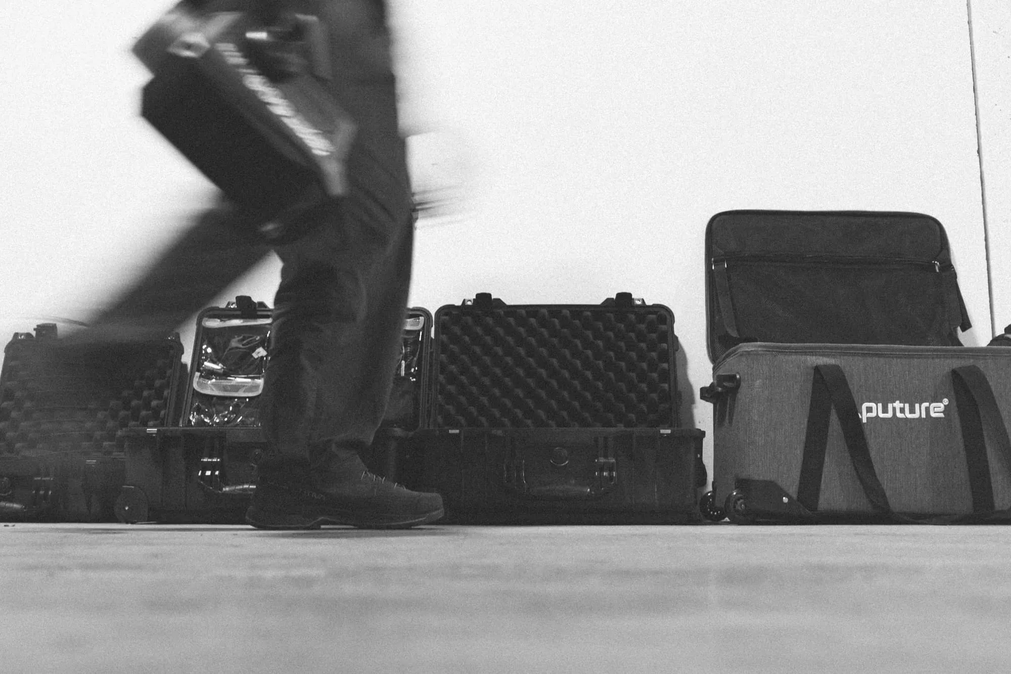 A black and white photo shows a person walking past several hard and soft cases lined up against a wall, including one labeled future. The person is carrying a case and only their legs are visible.