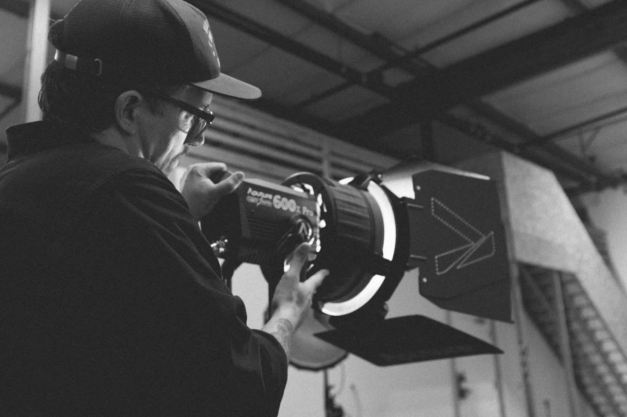 A person wearing glasses and a cap adjusts a large professional studio light labeled Aputure 600d Pro inside a warehouse or studio space. The image is in black and white.