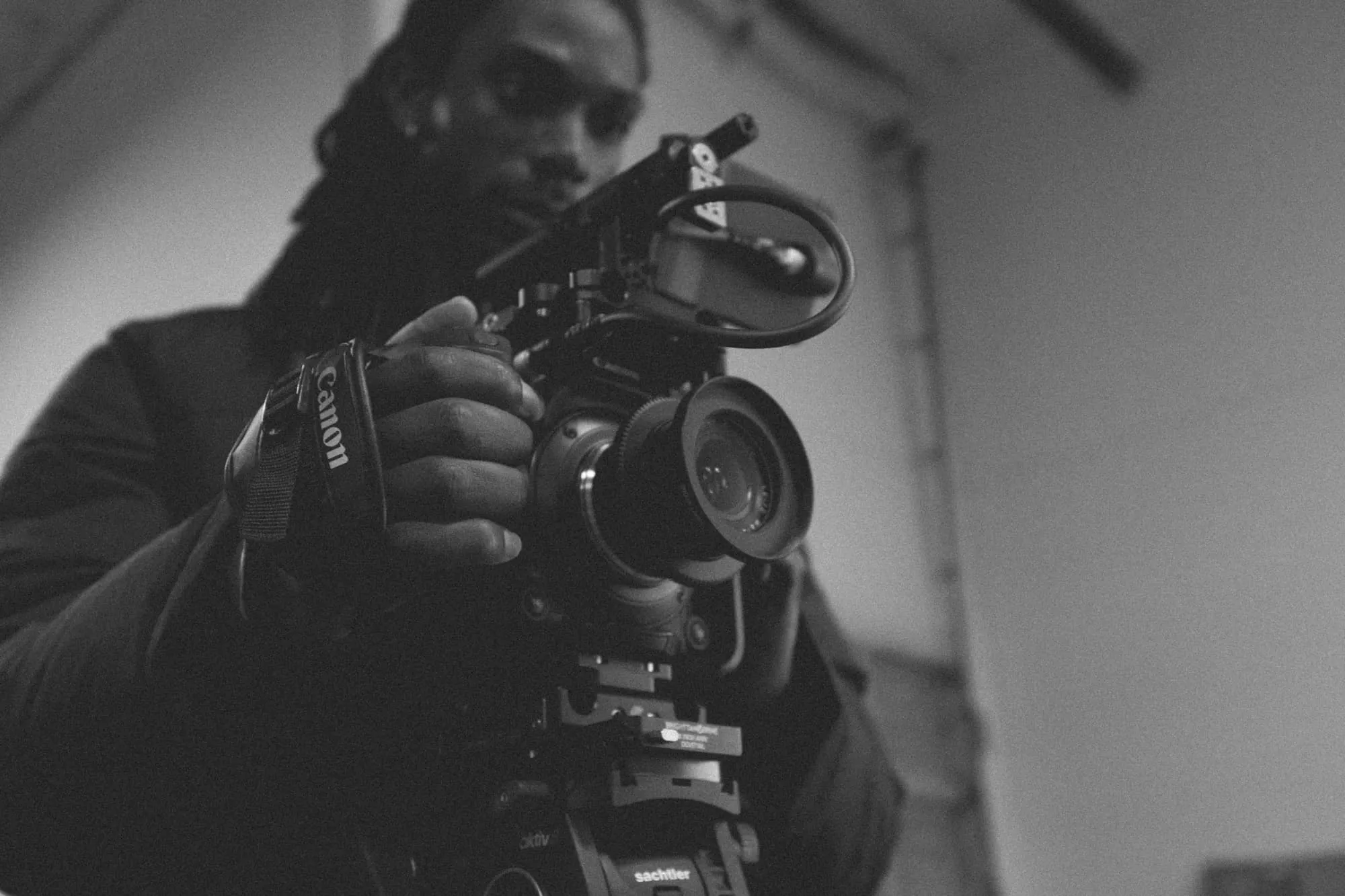 A person holding and adjusting a professional video camera, shot in black and white, with the camera lens prominently in the foreground and the person slightly out of focus in the background.