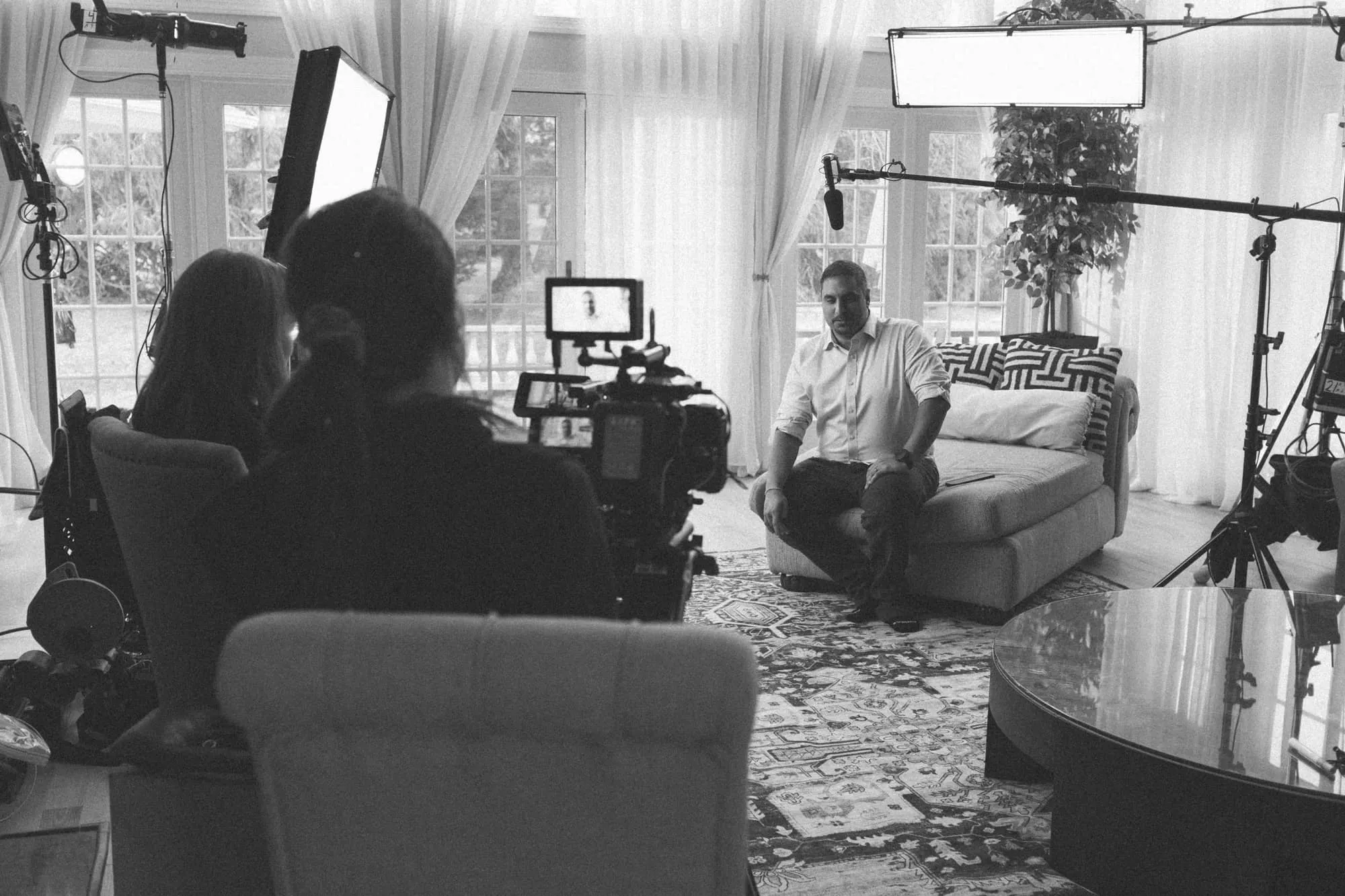 A black and white photo of a man sitting on a couch during a filmed interview, with cameras, microphones, and lighting equipment around him in a bright room with large windows and sheer curtains.