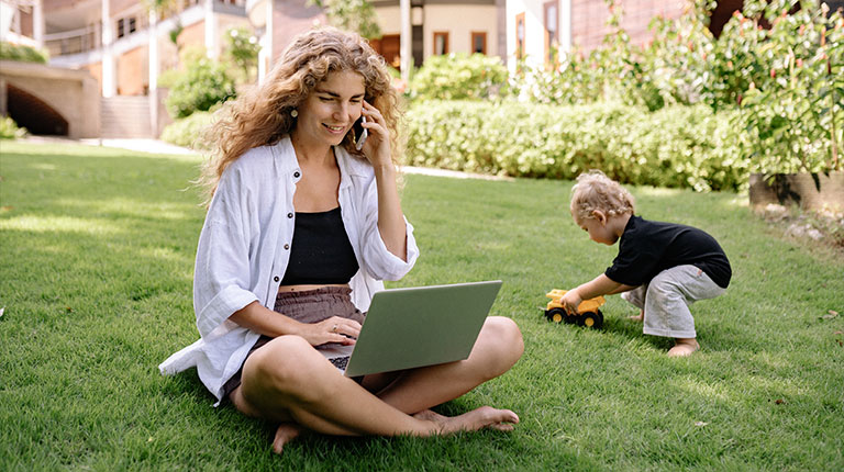 A woman using her phone and laptop, while her child plays in the background
