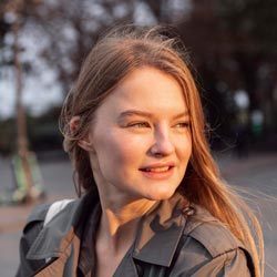 A person with long hair smiles while looking away, standing outdoors in casual clothing.