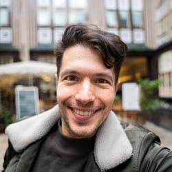 A person with short dark hair smiles for a selfie outdoors in front of a building.
