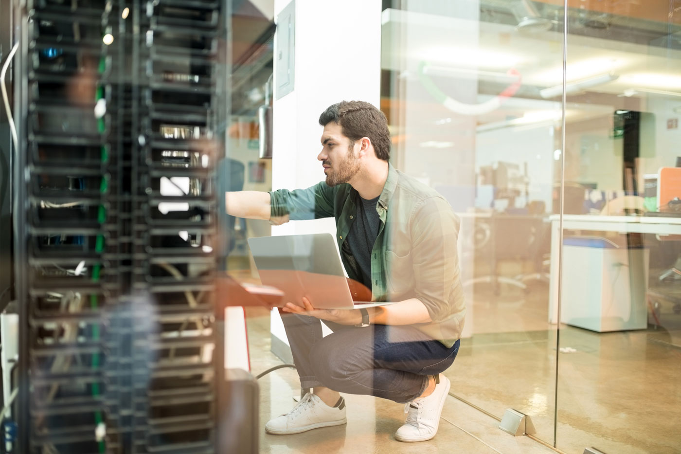 A man kneels while working on servers, holding a laptop in an office setting.