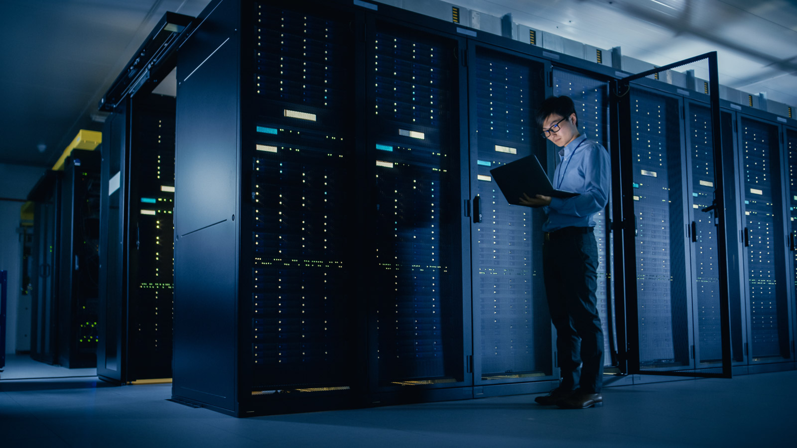 A person stands in a server room, reading from a laptop.