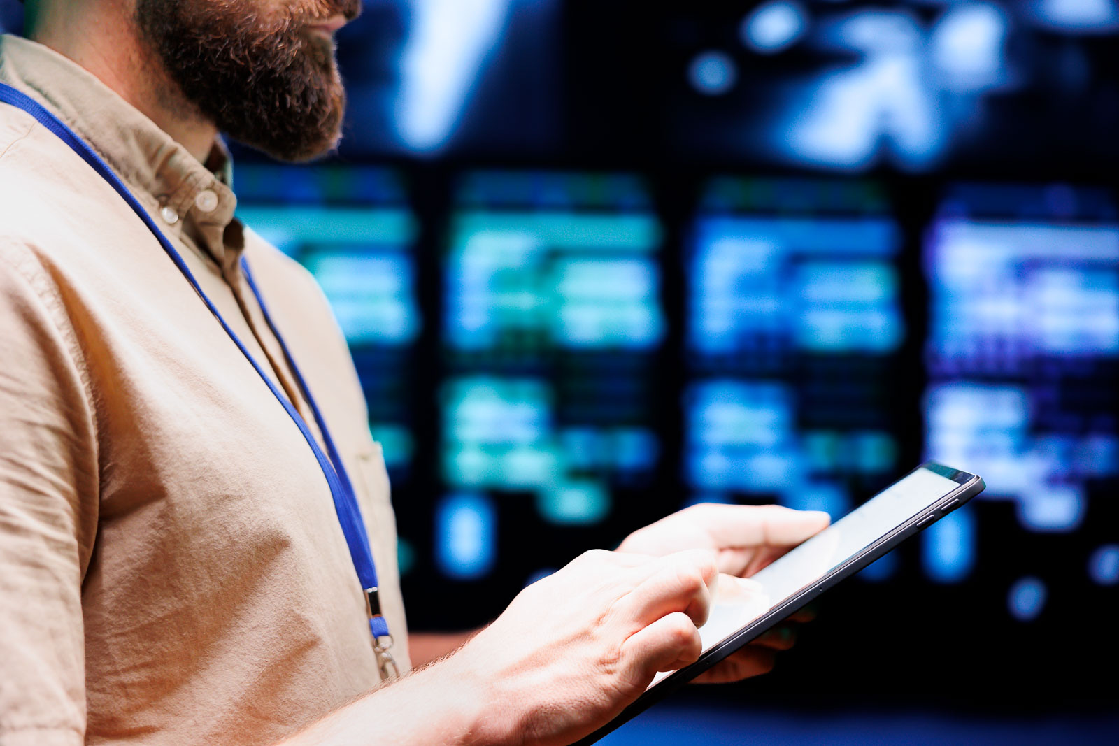 Man with a beard using a tablet in front of large data screens.