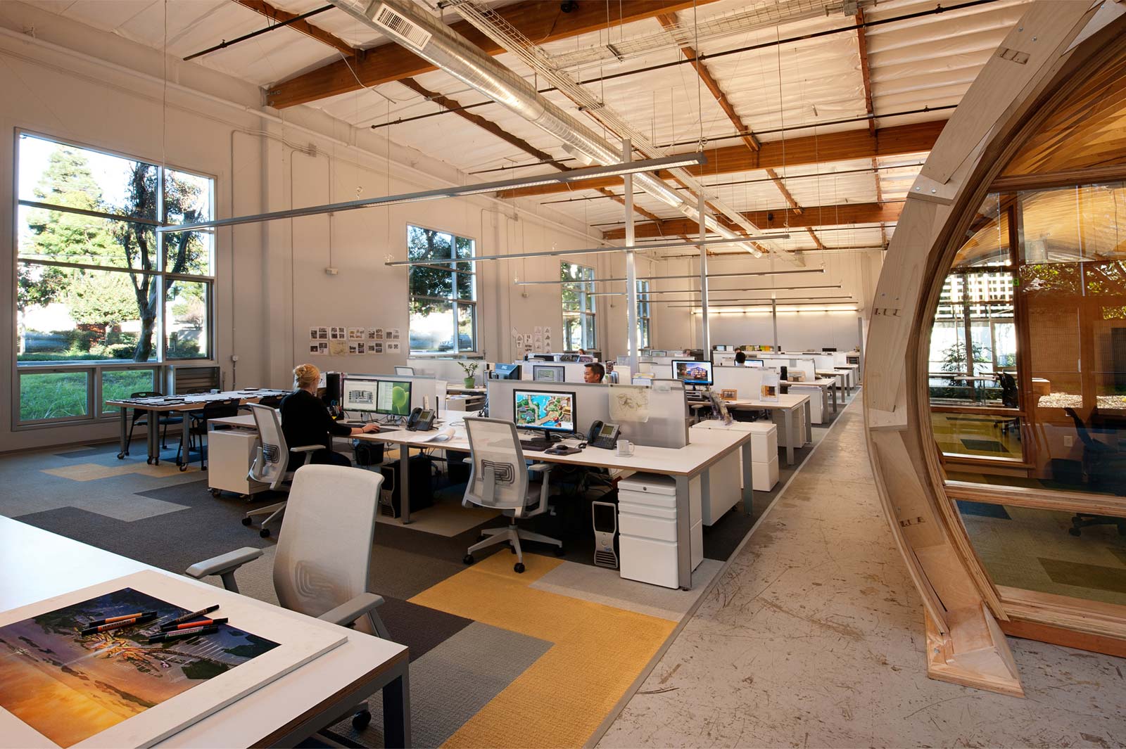 An office with open workstations, occupied by people working on computers. Large windows let in natural light.