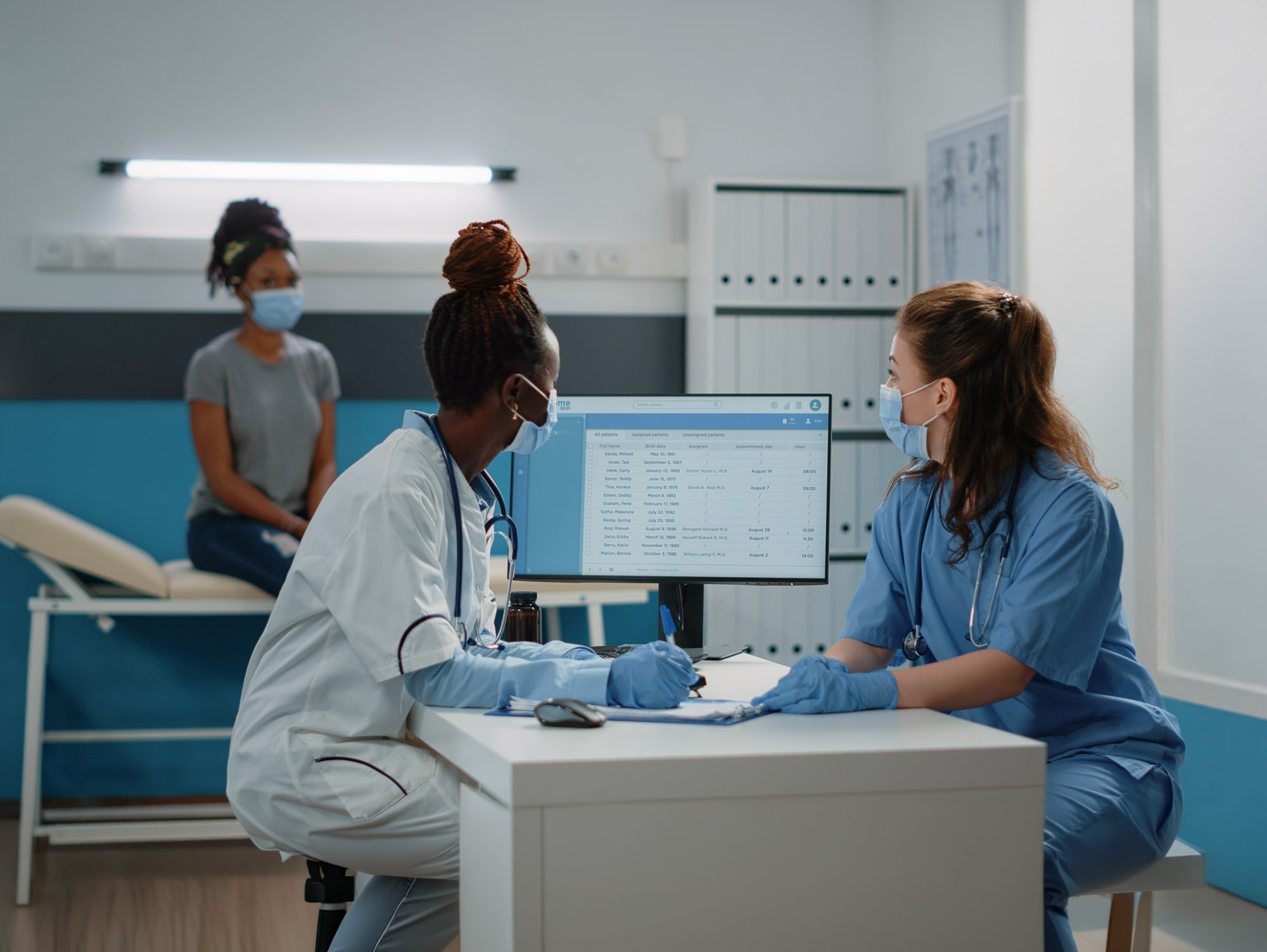 Two healthcare workers discuss patient information on a computer while a patient waits in the background.