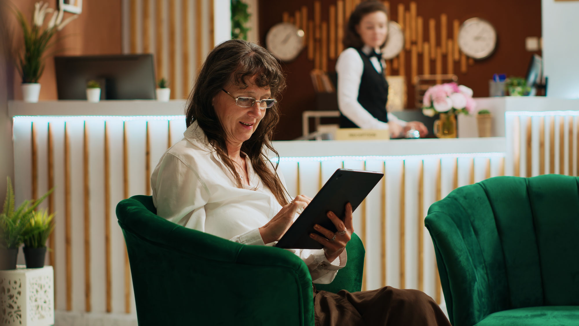 Woman sitting in green chair, writing on a clipboard, with a receptionist in the background.