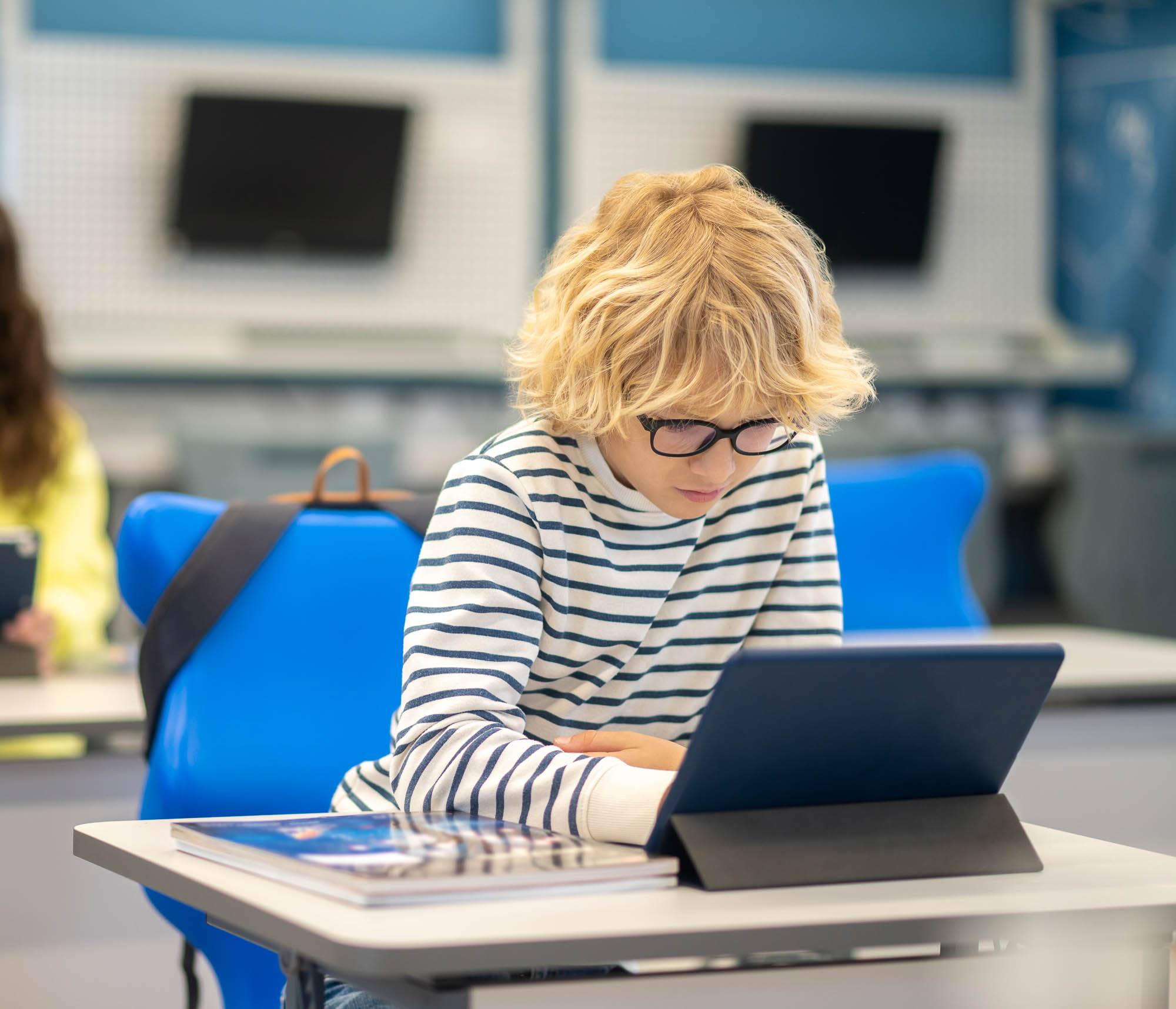 A child with glasses focuses on a tablet while sitting at a school desk.