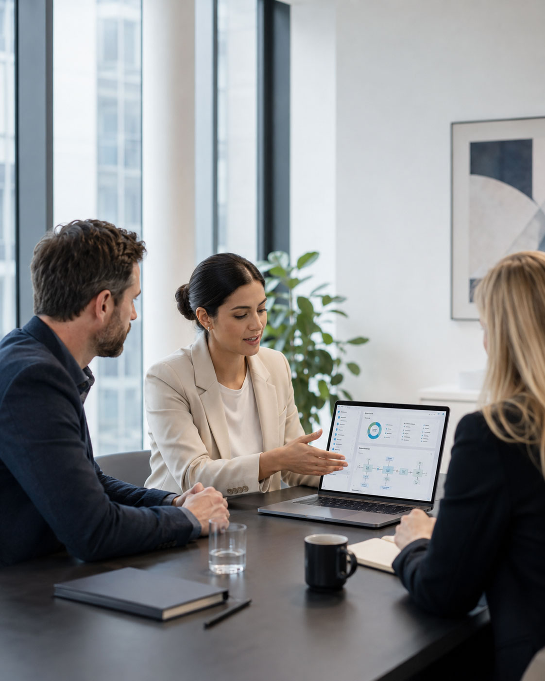 A woman in a suit presents data on a laptop to two colleagues at a conference table.