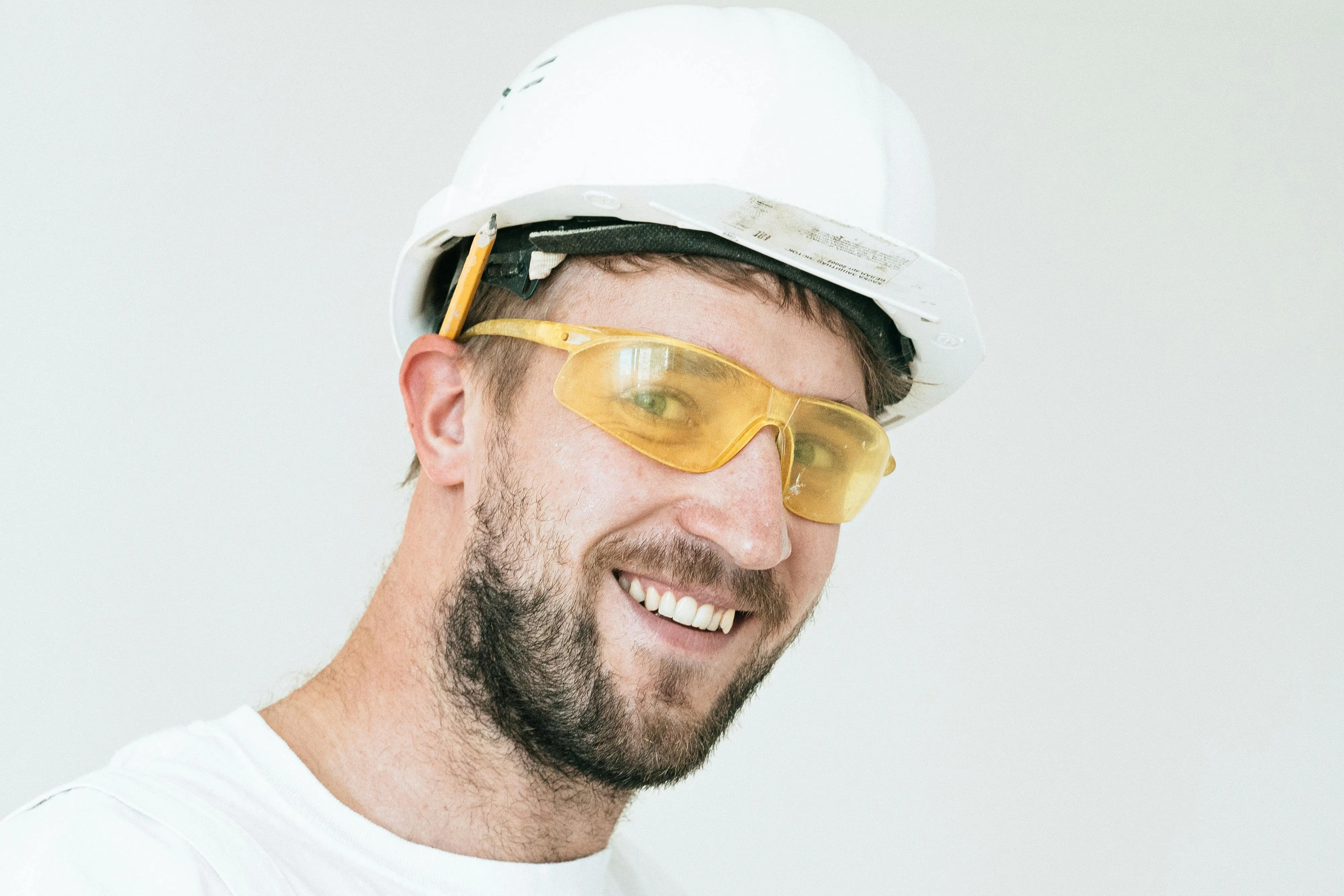 Man wearing a white construction helmet with a pencil tucked in and yellow safety glasses smiling.
