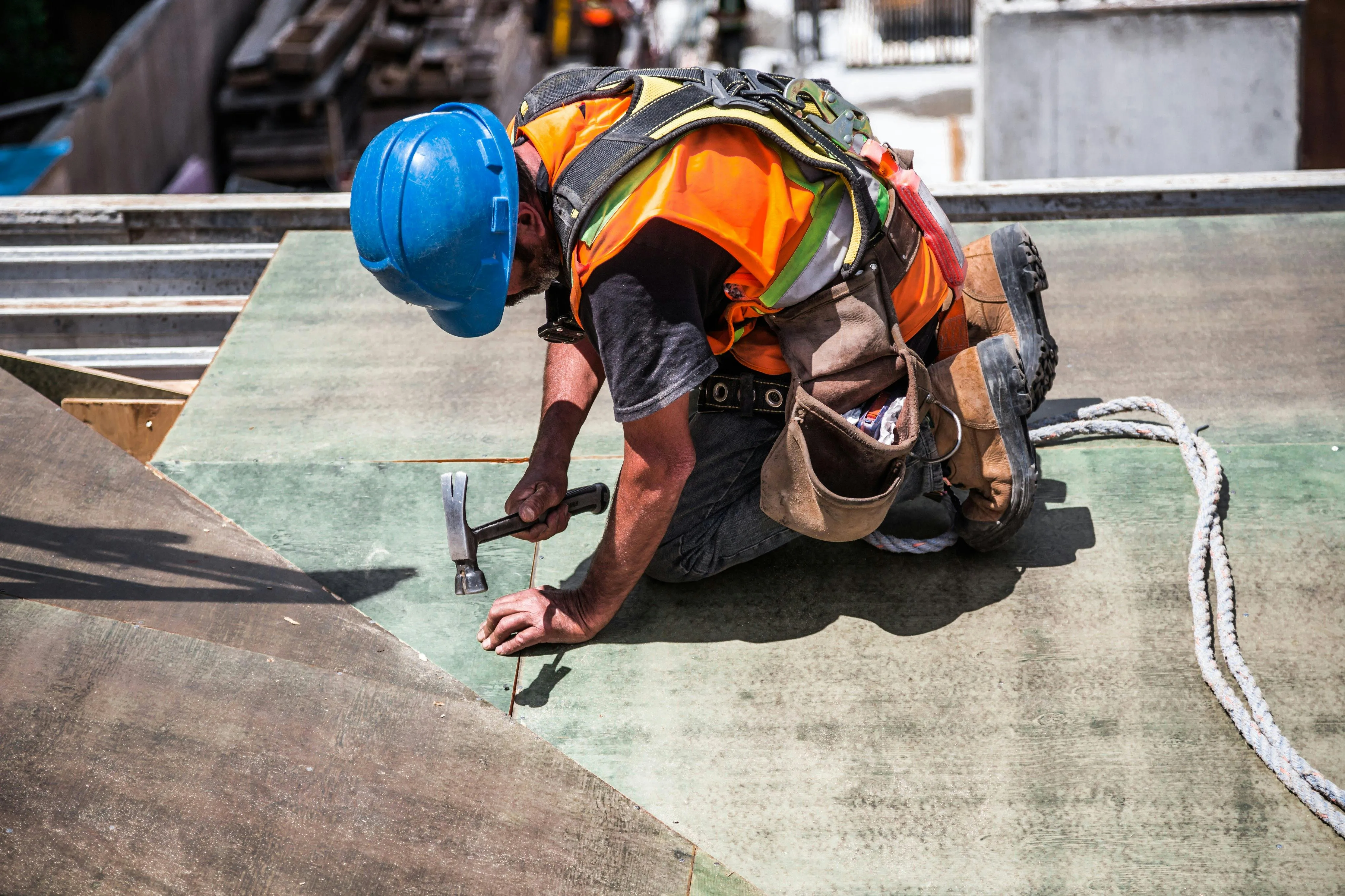 Construction worker wearing a blue hard hat and orange vest kneeling while hammering a nail into a wooden surface.