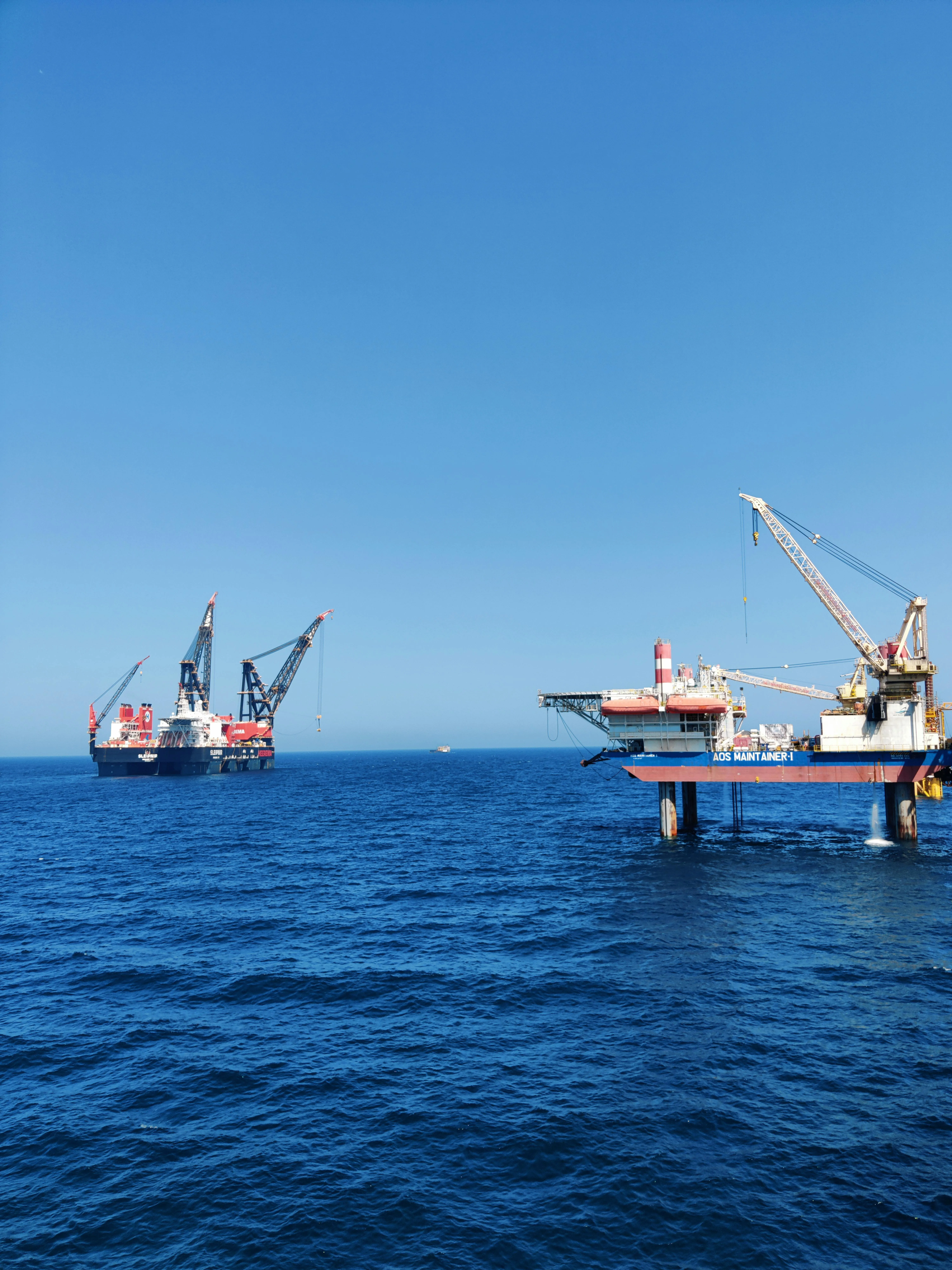 Offshore oil platform and a large crane ship on calm blue ocean under a clear sky.