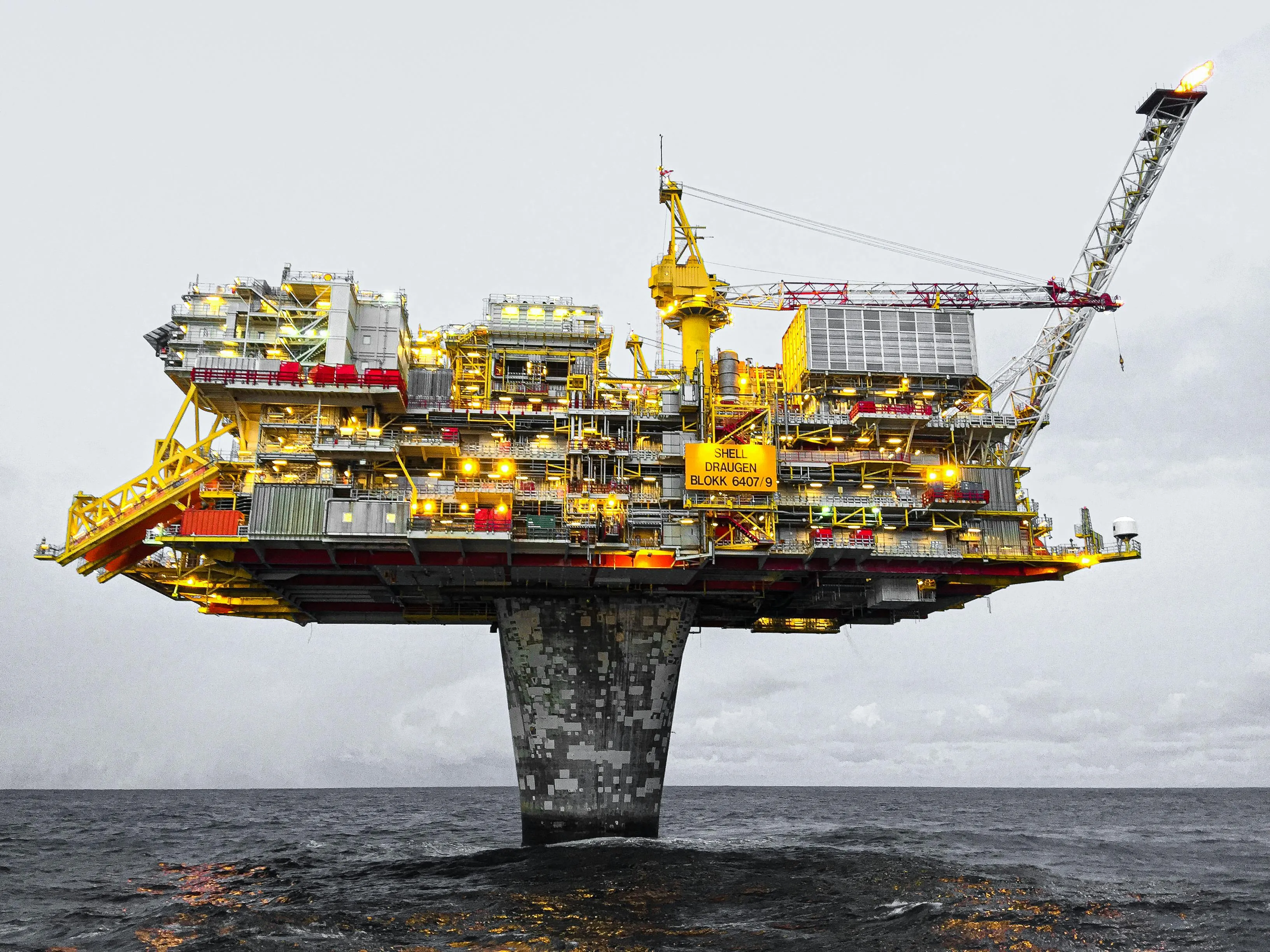 Illuminated Shell Draugen offshore oil platform on a single concrete support in the sea during cloudy weather.