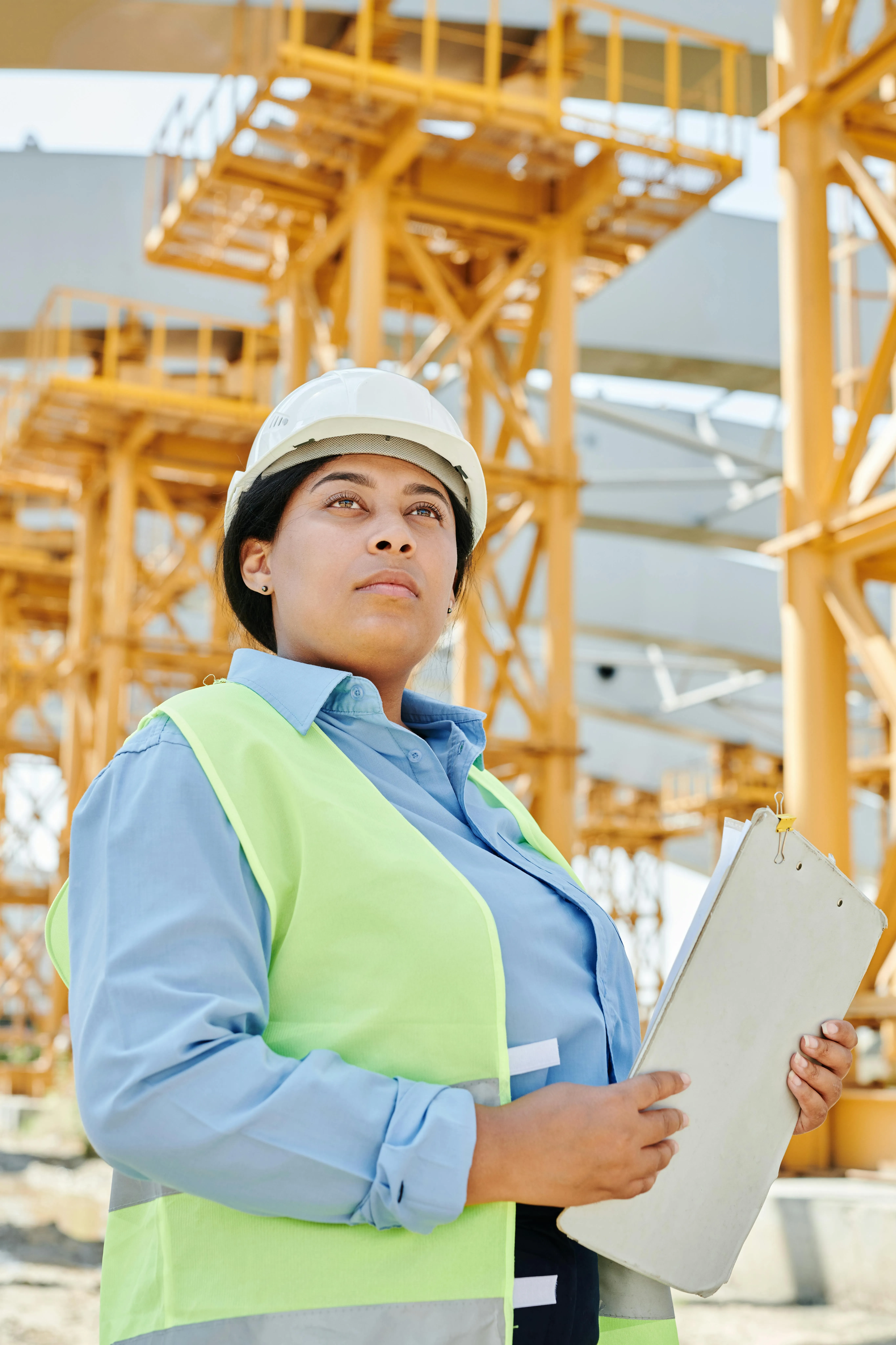 Woman construction engineer wearing a hard hat and safety vest holding a clipboard at a construction site with yellow structural framework.