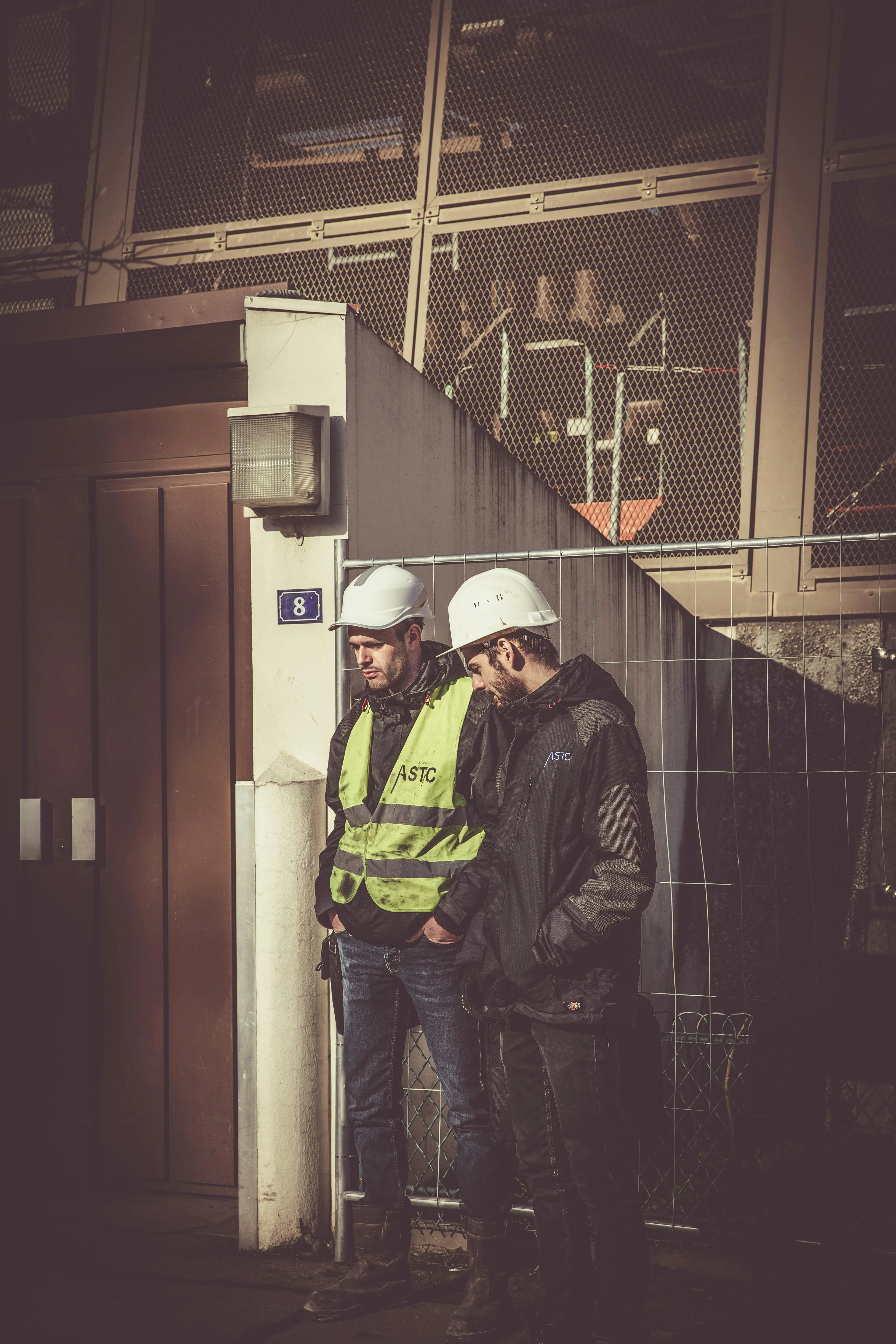 Two construction workers wearing white hard hats and jackets standing beside a building with a metal fence.